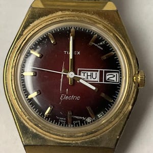 Könnte beinhalten: Eine goldfarbene Timex Electric Armbanduhr mit einem runden, dunkelroten Farbverlaufszifferblatt, goldenen Stundenmarkierungen und Zeigern. Das Zifferblatt zeigt den Text Timex, Electric und Taiwan sowie eine Datumsanzeige mit THU 21.