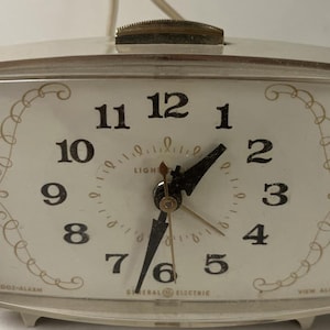 May include: A vintage General Electric electric alarm clock with a rectangular off-white plastic casing. The clock face features black Arabic numerals, gold decorative scrollwork, and black hour and minute hands. The text reads SNOOZE-ALARM and VIEW ALARM.