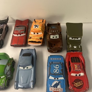 May include: A collection of die-cast toy cars featuring various characters with painted eyes on the windshields. The set includes a red car with number 95, a blue car with number 86 and Dinoco text, a brown truck, an orange car with number 91, and others.
