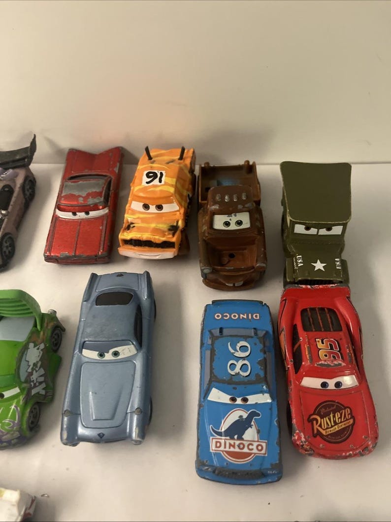 May include: A collection of die-cast toy cars featuring various characters with painted eyes on the windshields. The set includes a red car with number 95, a blue car with number 86 and Dinoco text, a brown truck, an orange car with number 91, and others.