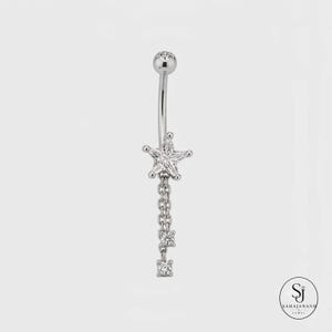 May include: A silver-toned curved barbell belly ring featuring a star-shaped charm set with clear stones. Two delicate silver chains hang from the star, each ending in a small, round, clear stone accent. The piece has a polished metallic finish.