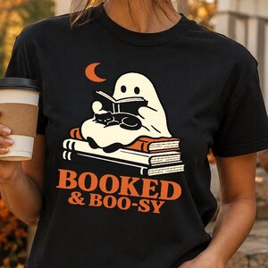 Può includere: Una t-shirt nera a maniche corte con la grafica di un fantasma bianco che legge un libro insieme a un gatto nero, seduto su una pila di libri sotto una luna crescente arancione. Il testo "BOOKED & BOO-SY" è stampato in arancione sotto.