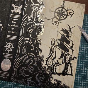 May include: A black leather book cover featuring silver foil stamping of a skull and crossbones, a ship's wheel, and the text "The republic of pirates" on the spine. The cover displays a large silver wave and a black ink illustration of a ship.