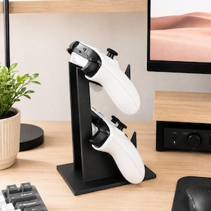 Op de afbeelding: Een zwarte verticale standaard ontworpen om twee gamecontrollers vast te houden. De standaard heeft een platte rechthoekige basis en twee trapsgewijze sleuven die witte draadloze controllers met zwarte joysticks, knoppen en triggers stevig ondersteunen.