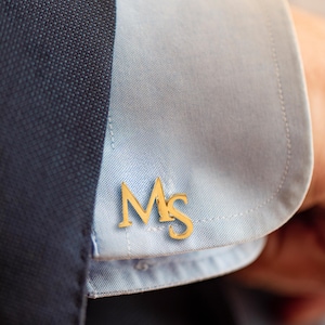 Op de afbeelding: Een goudkleurige metalen manchetknop met de letters M en S in een schreeflettertype. De manchetknop is bevestigd aan een lichtblauw overhemdmanchet, dat gedeeltelijk wordt bedekt door een donkerblauwe mouw van een jasje.