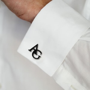 May include: A black metal cufflink featuring the letters AG in a serif font, attached to the white fabric cuff of a long-sleeved shirt. The letters are connected with the A positioned slightly above and to the left of the letter G.