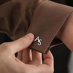 Op de afbeelding: Een bruine overhemdmouw met een zilverkleurige manchetknop in de vorm van de letters AS. De manchetknop is bevestigd aan de stoffen manchet, die een gladde, matte textuur heeft en zichtbare stiksels langs de randen van de mouwopening.