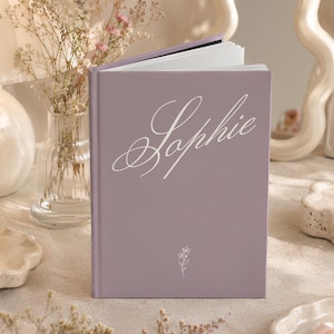 May include: A light purple hardcover book featuring the name Sophie in white cursive script on the front cover, accompanied by a small, delicate white line drawing of a flower centered below the name. The book is standing upright with white pages.