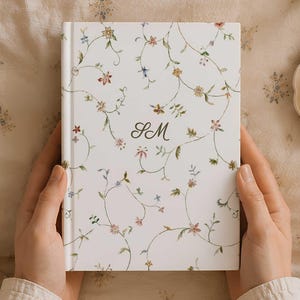 May include: A white hardcover journal featuring a delicate, repeating pattern of thin green vines and small, multicolored flowers. The center of the front cover displays the monogram letters SM in a dark, elegant, cursive script font.