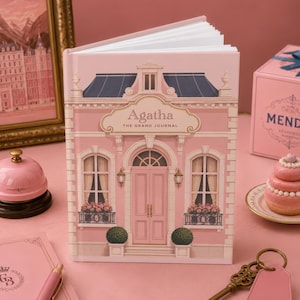 May include: A pink hardcover journal titled Agatha The Grand Journal, featuring a building facade illustration. The set includes a pink desk bell, a pink Mendl's box, a pink pastry, a pink notebook with a pen, and a brass key with a tag labeled 316.
