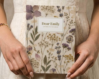 Può includere: Un libro rettangolare con copertina color crema caratterizzato da un motivo di fiori di campo pressati viola, gialli e verdi. Un'etichetta rettangolare bianca centrale riporta il testo Dear Emily LETTERS TO THE BRIDE in lettere dorate.