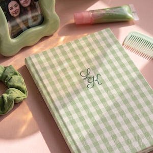 May include: A rectangular notebook with a light green and white gingham pattern cover featuring the embroidered monogram letters LGK in dark green thread, accompanied by a green scrunchie, a light green wavy picture frame, and a hair comb.