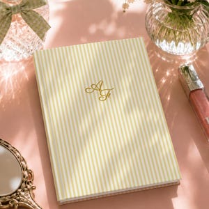 May include: A rectangular notebook featuring a vertical yellow and white striped pattern on the cover. The center of the cover displays the gold-colored monogram letters AF in a cursive script font. The notebook has a clean, hardbound exterior.