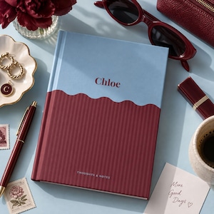 May include: A hardcover notebook featuring a two-tone design with a light blue top section and a dark red bottom section with vertical stripes. The name Chloe is printed in dark red on the top, and the text THOUGHTS & NOTES is at the bottom.