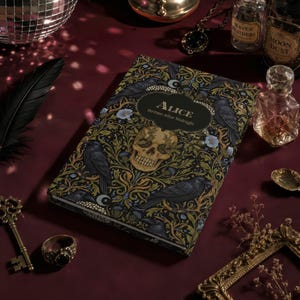 Pu&ograve; includere: Un libro con copertina rigida rettangolare intitolato Alice Written After Midnight, caratterizzato da un motivo botanico scuro con corvi neri, fiori blu e un'illustrazione centrale di un teschio umano su sfondo nero con accenti oro e verdi.