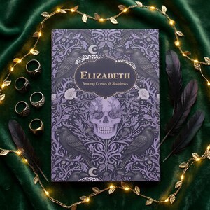 Pu&ograve; includere: Un libro rettangolare intitolato Elizabeth Among Crows & Shadows con una copertina viola raffigurante un teschio, corvi e motivi floreali. Cinque anelli in metallo sono disposti a sinistra e due piume nere a destra del libro.