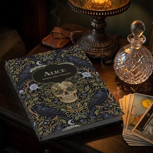 Pu&ograve; includere: Un libro con copertina rigida intitolato Alice Written After Midnight caratterizzato da un motivo botanico scuro con corvi neri, fiori bianchi e un teschio umano centrale. Il libro &egrave; posizionato accanto a un decanter in cristallo e tarocchi.