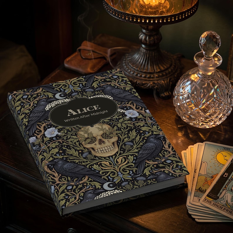 Pu&ograve; includere: Un libro con copertina rigida intitolato Alice Written After Midnight caratterizzato da un motivo botanico scuro con corvi neri, fiori bianchi e un teschio umano centrale. Il libro &egrave; posizionato accanto a un decanter in cristallo e tarocchi.