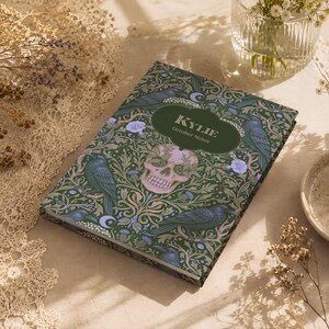 Pu&ograve; includere: Un taccuino rettangolare con copertina verde scuro caratterizzato da un motivo ripetuto di corvi neri, fiori viola chiaro e un teschio umano centrale. Un'etichetta ovale verde scuro riporta il testo bianco "KYLIE" e "October Notes".