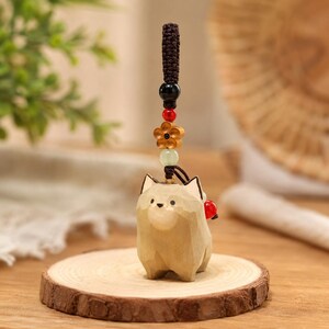 May include: A hand-carved wooden dog figurine keychain featuring a light brown finish with faceted surfaces. The charm is attached to a dark brown braided cord adorned with a black bead, a red bead, a light wood flower, and a pale green bead.