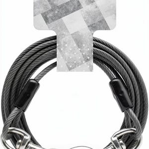 Puede incluir: Una correa de cable de acero gris enrollado con un revestimiento de plástico protector, dos ganchos de seguridad giratorios de metal plateado en los extremos y conectores engarzados de plástico negro, fijada a un encabezado de cartón gris y blanco.