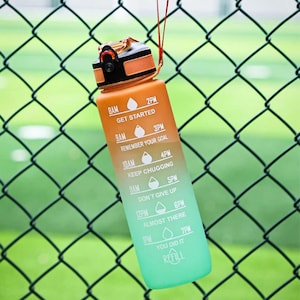May include: A plastic water bottle with an orange-to-green gradient finish, featuring a black and orange lid with a carrying strap. The bottle displays white time markers and motivational text including GET STARTED, REMEMBER YOUR GOAL, KEEP CHUGGING, DON'T GIVE UP, ALMOST THERE, YOU DID IT, and REFILL.