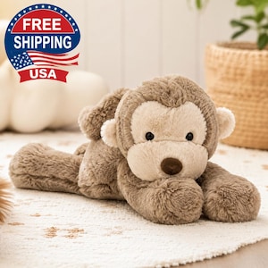 May include: A plush monkey toy with soft, light brown fur and a cream-colored face. The stuffed animal features small round ears, a dark brown nose, and black bead eyes. It is positioned in a lying down pose with its limbs extended forward.