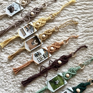 Può includere: Una collezione di sette decorazioni da parete in macramè in vari colori, tra cui crema, marrone, giallo, arancione, marrone scuro, verde salvia e verde scuro. Ogni pezzo presenta un anello in legno, corda annodata, una perlina e una foto.