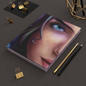 Könnte beinhalten: Ein rechteckiges Notizbuch mit einer detaillierten digitalen Illustration eines Gesichts mit einem markanten blauen Auge, dunklem Haar mit türkisfarbenen Highlights und rosa Lippen. Das Cover zeigt das Wort "Vanderon" in dezenter Schreibschrift.