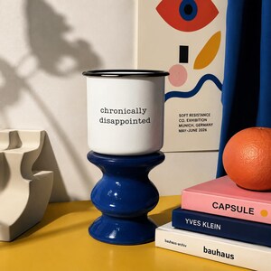 May include: A white enamel mug with a black rim and the text "chronically disappointed" printed in black, resting on a glossy, dark blue, hourglass-shaped pedestal. The mug is positioned in front of a poster and a stack of three books.