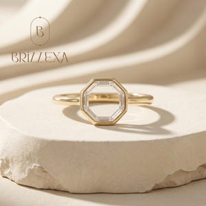 Peut inclure: Une bague en or avec un sertissage octogonal composé de pierres transparentes taille baguette. L'anneau est lisse et poli. Le texte "BRILLEXA" et un logo avec la lettre "B" sont visibles dans le coin supérieur gauche de l'image.