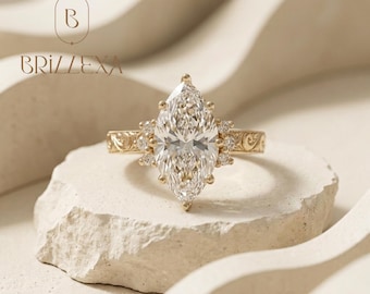 May include: A gold ring featuring a large marquise-cut centre stone flanked by smaller round stones on each side. The band displays an intricate engraved scrollwork pattern. The text BRIZLEXA and a logo with the letter B are visible above the ring.