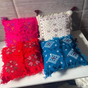 Peut inclure: Quatre coussins carrés ornés de broderies décoratives et de franges sur les bords. L'ensemble comprend un coussin rose vif, un rouge, un blanc et un bleu, chacun décoré de motifs géométriques et de petits accents métalliques sur le tissu.