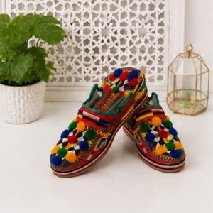 May include: A pair of slip-on shoes featuring a vibrant multi-colored geometric pattern, accented with numerous small, fuzzy pom-poms in shades of red, blue, green, and yellow, finished with a thick, layered sole and green trim along the collar.