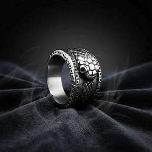 May include: A silver-toned metal ring featuring a detailed snake head design on the outer band. The surface of the ring displays a textured scale pattern across the snake's head and body, with a smooth, polished interior band for a comfortable fit.
