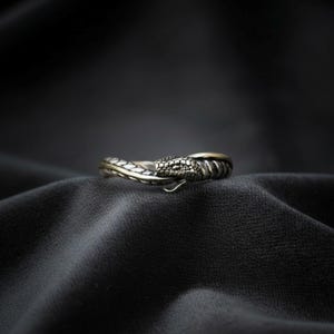 Puede incluir: Un anillo plateado con forma de serpiente enroscada con escamas detalladas en la cabeza y el cuerpo. El metal tiene un acabado oscuro y oxidado en las zonas hundidas, resaltando la textura del diseño de serpiente que rodea el dedo.