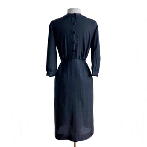 May include: A dark blue, knee-length dress with a fitted bodice and a slightly flared skirt. The dress has a round neckline and long sleeves with button cuffs. There are five buttons down the back of the dress.