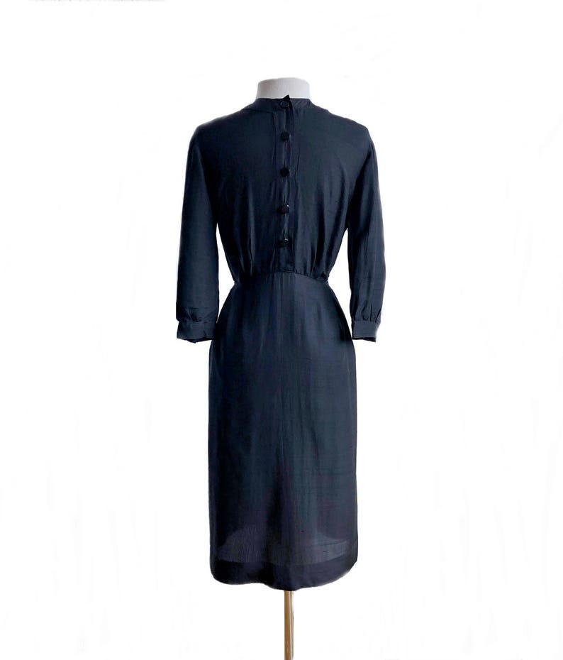 May include: A dark blue, knee-length dress with a fitted bodice and a slightly flared skirt. The dress has a round neckline and long sleeves with button cuffs. There are five buttons down the back of the dress.