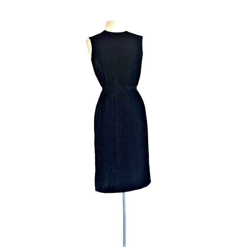 May include: A sleeveless black dress with a fitted bodice and a straight skirt. The dress is made of a textured fabric and has a simple, elegant design.