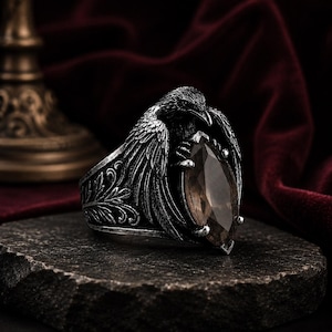 May include: A silver-toned ring featuring a detailed raven with spread wings clutching a marquise-cut, translucent brown gemstone. The band displays intricate, embossed leaf-like patterns along the sides, with a dark, oxidized finish.