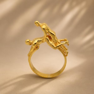 Puede incluir: Un anillo de tono dorado con un diseño escultórico de dos figuras en una pose entrelazada. La banda es circular y lisa en el interior, mientras que la superficie exterior muestra un acabado metálico texturizado sobre las dos formas humanas.