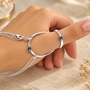 May include: A silver hand chain featuring a delicate double chain bracelet connected to an oval-shaped frame that encircles the thumb. The piece is accented with two small, round, deep blue gemstones set into the silver metal frame and ring.