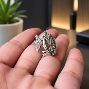 May include: A silver-toned metal ring featuring a detailed dragon design with spread wings, a long curved tail, and textured scales. The dragon is positioned centrally on the band, showcasing an oxidized finish that highlights the intricate carvings.