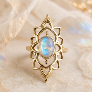 Peut inclure: Une bague dorée ornée d'une pierre de lune ovale et iridescente en son centre. La pierre est entourée d'une monture métallique symétrique et complexe, dotée de découpes pointues en forme de pétales s'étendant verticalement autour du bijou.