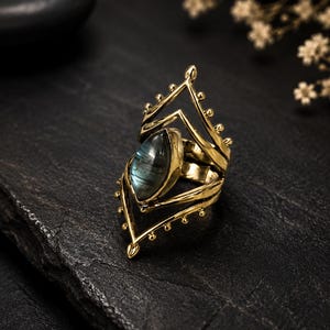 May include: A gold-toned metal ring featuring a marquise-cut labradorite gemstone at its centre. The band is designed with an open, geometric frame that extends upwards and downwards, accented with small metal spheres along the outer edges.