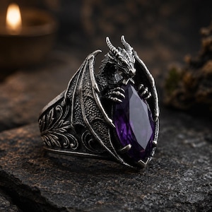 May include: A silver-toned ring featuring a sculpted dragon with spread wings holding a large, marquise-cut purple gemstone in its claws. The band of the ring is decorated with intricate, raised scrollwork patterns along the sides.