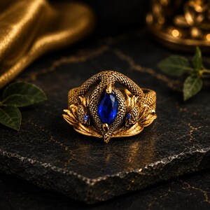 Puede incluir: Un anillo de tono dorado con un motivo de serpiente texturizada que envuelve una piedra preciosa azul central de corte marquesa. El diseño de la serpiente incluye escamas detalladas y pequeños acentos azules en las cabezas laterales.