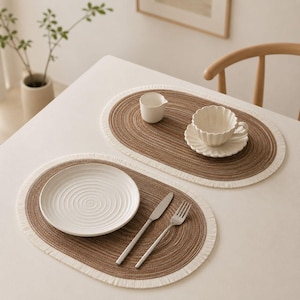 Puede incluir: Dos manteles individuales ovalados con centro tejido marrón y borde de flecos blanco. Uno sostiene un plato blanco, un cuchillo y un tenedor plateados. El otro sostiene una taza de té blanca festoneada sobre un plato y una pequeña jarra de leche.