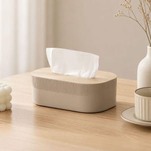 Può includere: Un porta scatola di fazzoletti di forma ovale con un coperchio color legno chiaro e una base beige con dettagli a coste verticali. Un singolo fazzoletto bianco è estratto attraverso l'apertura centrale del coperchio, sporgendo verso l'alto.