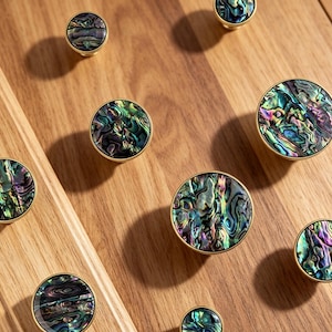 May include: A collection of round cabinet knobs featuring iridescent abalone shell inlays with swirling patterns of green, blue, and purple hues, each encased in a polished gold-toned metal rim, arranged in various sizes across the frame.